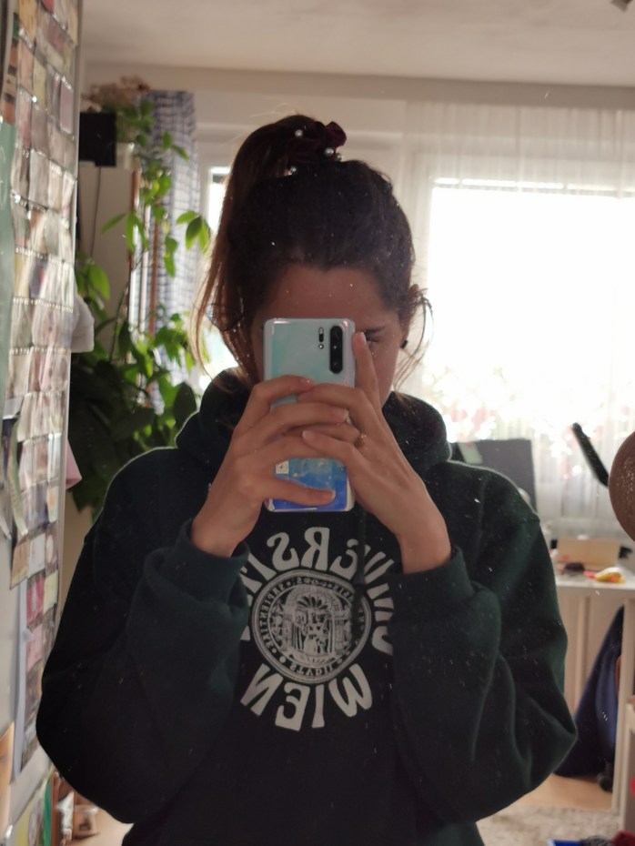 Frau fotografiert sich im Spiegel. Gesicht ist mit dem Handy verdeckt, am Pulli ist das Logo der Universität Wien.